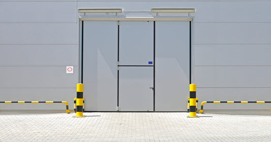 The outside door of a gray warehouse with yellow and black safety bollards and rails in front of it.