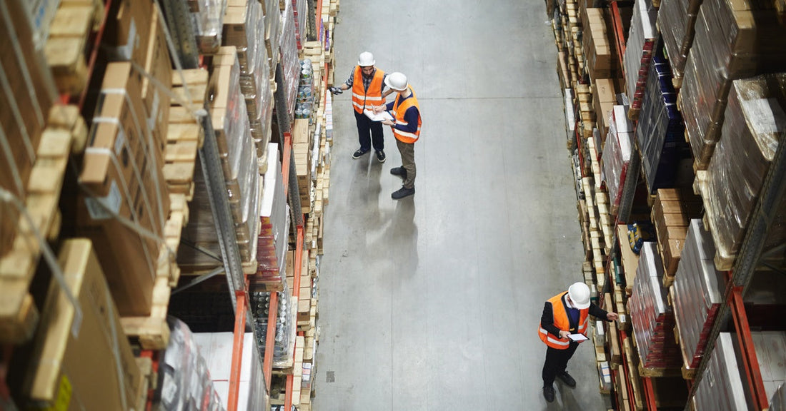 Warehouse workers in orange safety vests in an aisle between rows of tall shelves full of packed boxes and goods.