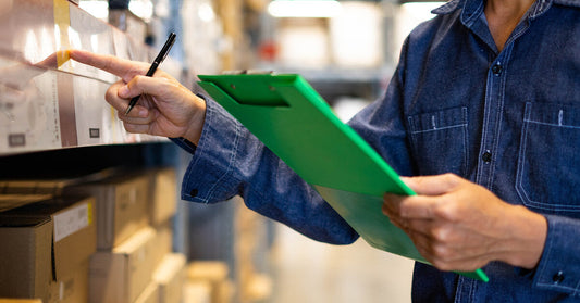 person holds a green clipboard in their left hand and a pen in their right hand while looking at goods on a shelf.