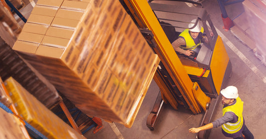 Two men in a warehouse wearing safety vests. One man stands and points, guiding the other man as he operates a machine.