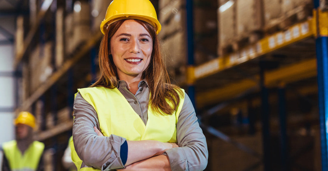 A woman wearing a yellow hard hat and safety vest smiles as she stands in a warehouse with her arms folded.