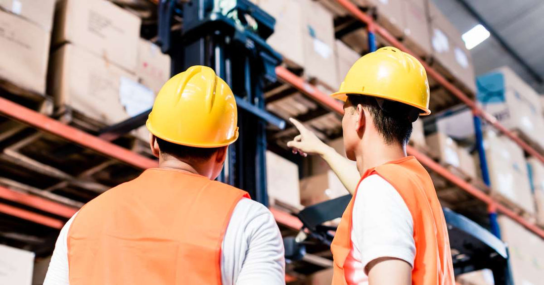 Two warehouse workers in yellow hard hats and orange safety jackets look up at a pallet racking system, with one pointing to a higher shelf.