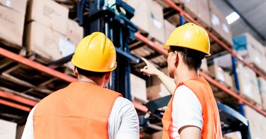 Two warehouse workers in yellow hard hats and orange safety jackets look up at a pallet racking system, with one pointing to a higher shelf.