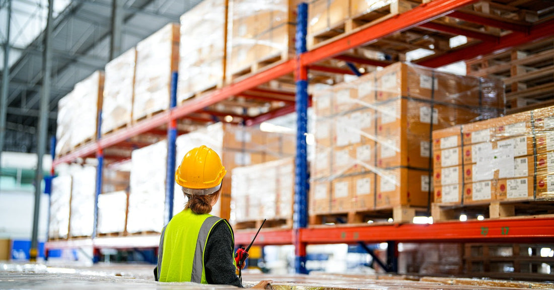 A worker wearing a hard hat and yellow safety vest checking labels on shrink-wrapped pallets in a warehouse.