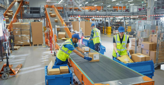 Three people in safety vests sorting cardboard boxes on a conveyor inside a warehouse with tall shelves.
