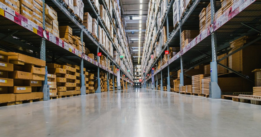 A wide warehouse aisle lined with tall metal racks filled with cardboard boxes and wrapped pallets under bright lights.