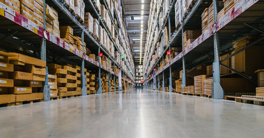 A wide warehouse aisle lined with tall metal racks filled with cardboard boxes and wrapped pallets under bright lights.