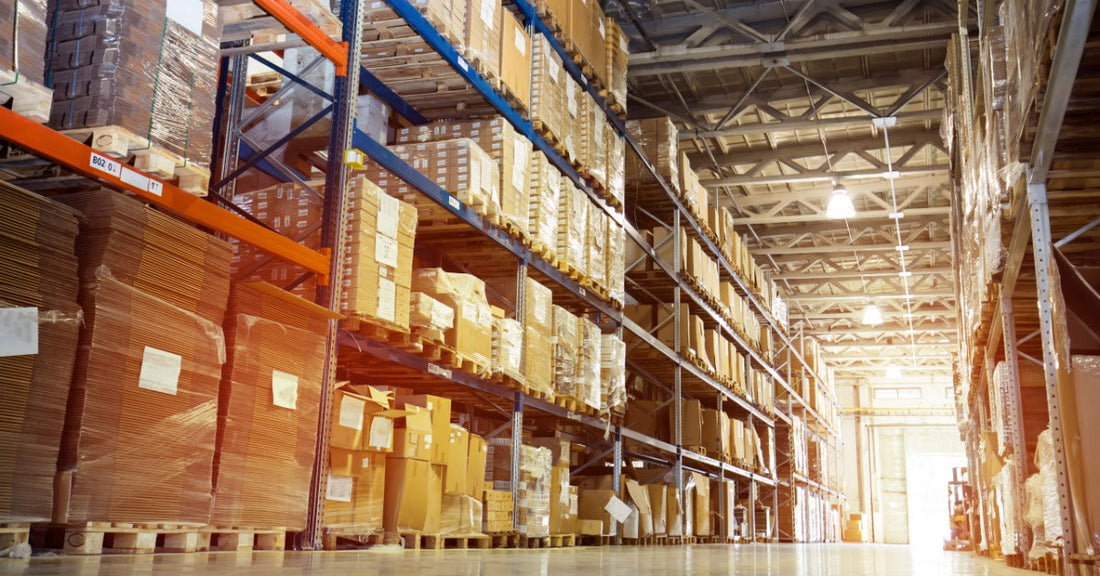 Tall warehouse pallet racks filled with stacked cardboard boxes and wrapped pallets line a wide aisle beneath a high ceiling.