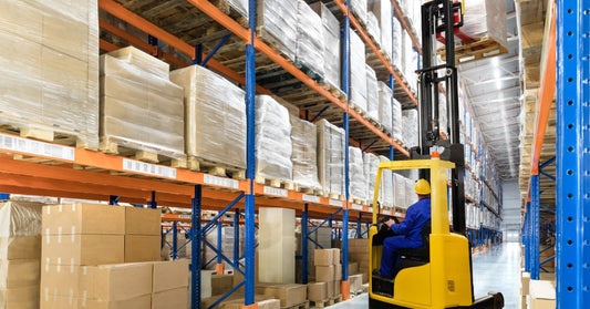 A worker in blue coveralls and a yellow hard hat operating a yellow forklift between tall racks of wrapped pallets.