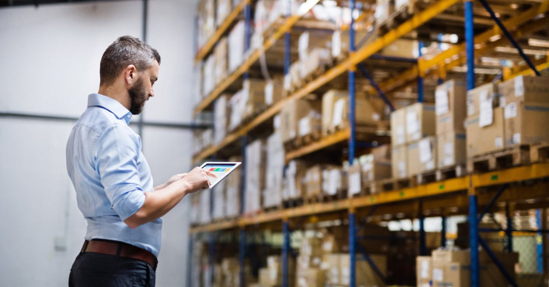 A warehouse worker is looking down at a tablet while standing in front of a large shelf of supplies.