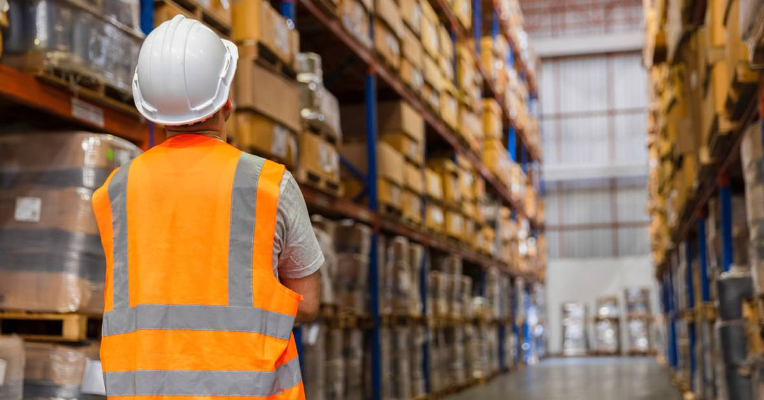 The back of person wearing a safety vest standing in front of pallet rack shelves full of cardboard boxes.