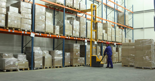 A man in a blue safety suit operates a machine in a warehouse filled with boxes stacked on pallet racks.