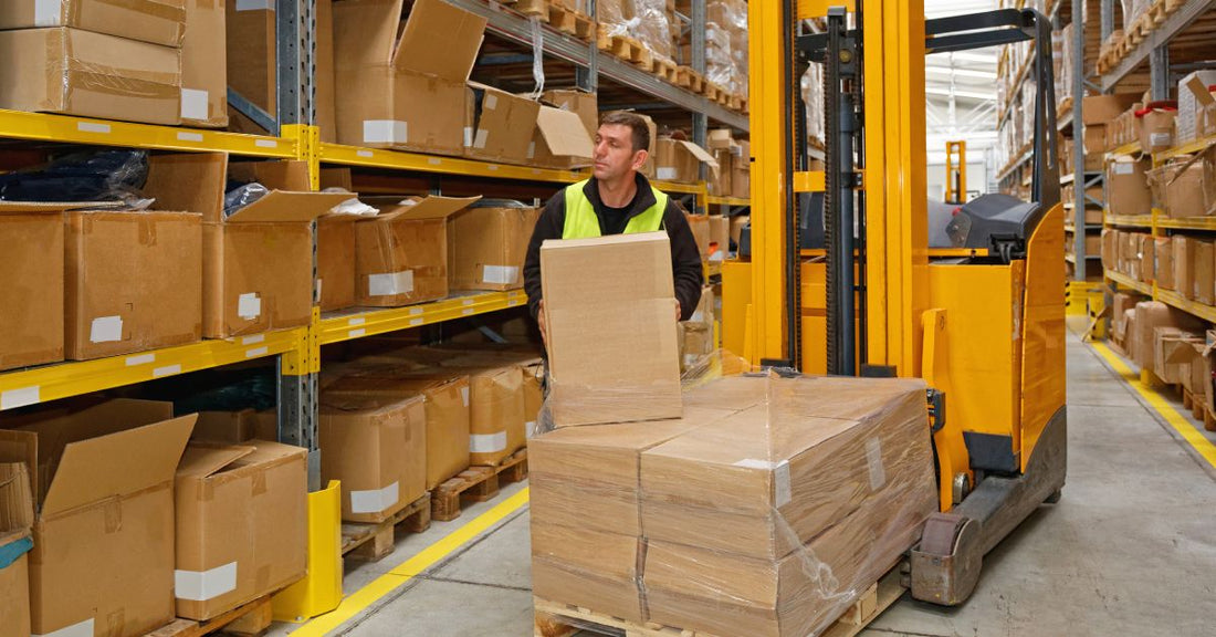 A man in a warehouse, wearing a safety vest, holds a brown cardboard box as he stands next to racks that store other boxes.