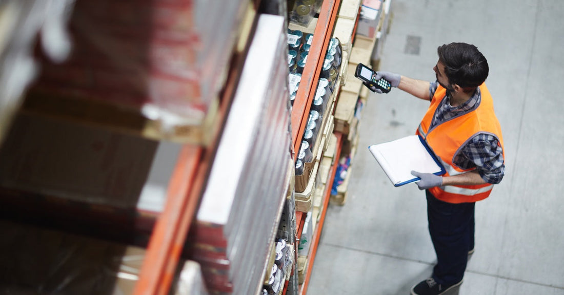 A worker walking around with a scanner to review goods, all stacked on a shelf. He is in a warehouse.