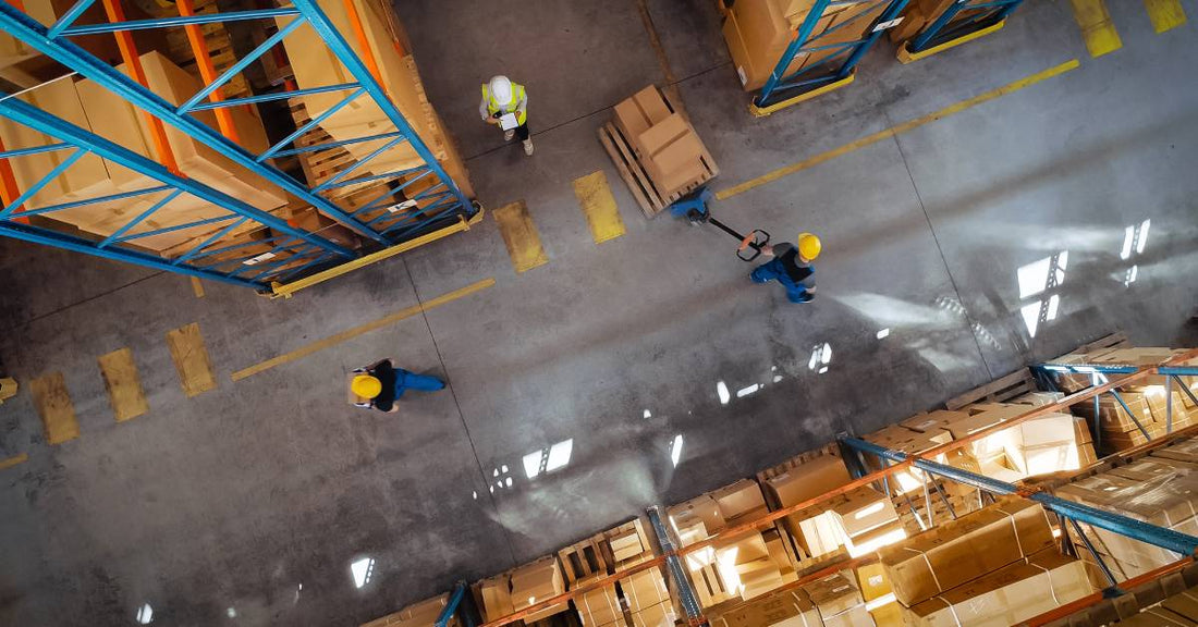 An overhead view of workers moving around a warehouse floor. One worker is pulling a large pallet of inventory on a jack.