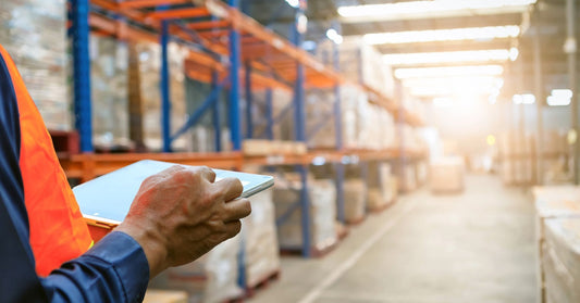A man standing in a warehouse. He is holding a tablet while checking observing the product stock on the shelves around him.