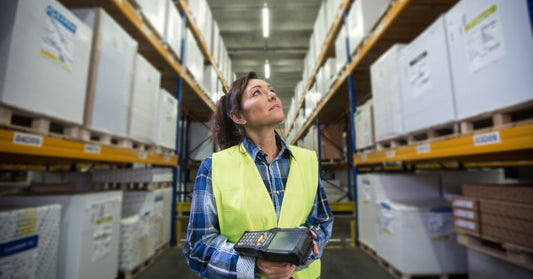 A woman with a hand scanner looking up at the warehouse shelves around her. There are multiple boxes on the racks.