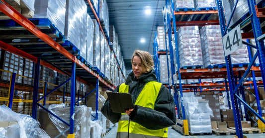 A worker in a cold storage area is checking inventory on a clipboard. There are large shelving racks around her.