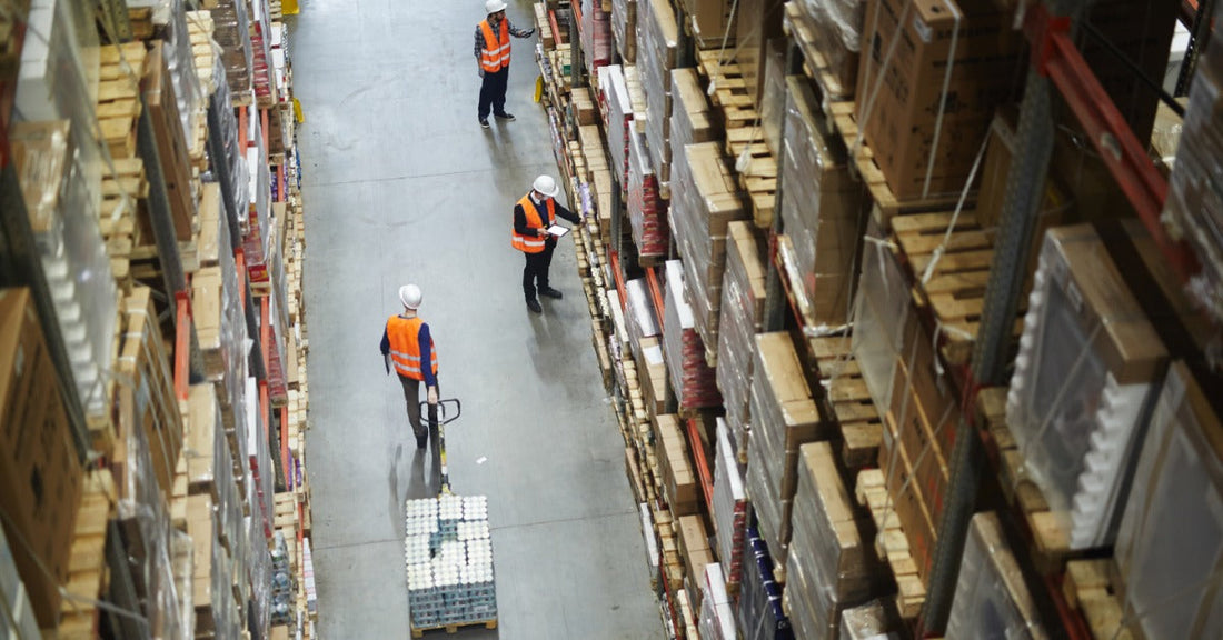 A warehouse with multiple workers moving through a row of large pallet racking units. One worker is pulling a pallet.