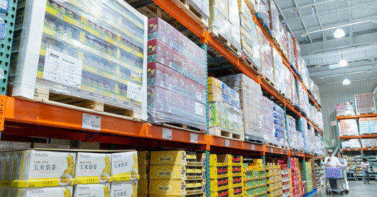 A warehouse with pallet racking shelves holding a variety of shopping items. Down the row, there are people shopping.