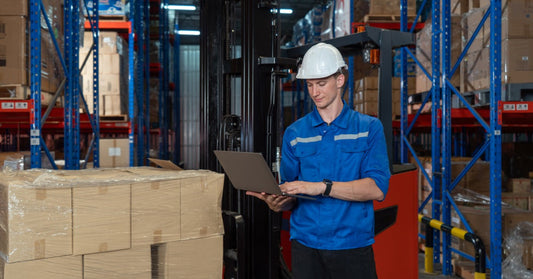A worker in a warehouse with safety gear on. He is looking at a laptop with shelving and a forklift behind him.