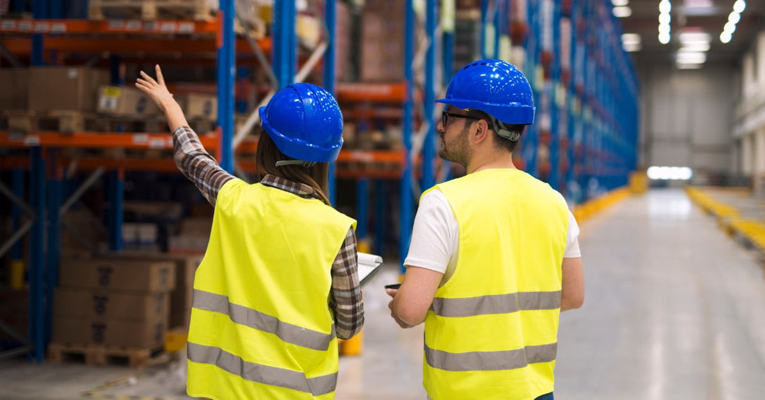 Two warehouse workers in hard hats walking with a clipboard past aisles of pallet racks while speaking to each other.