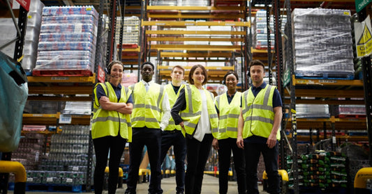 A group of employees in a warehouse standing together. They are surrounded by industrial shelves with packages.