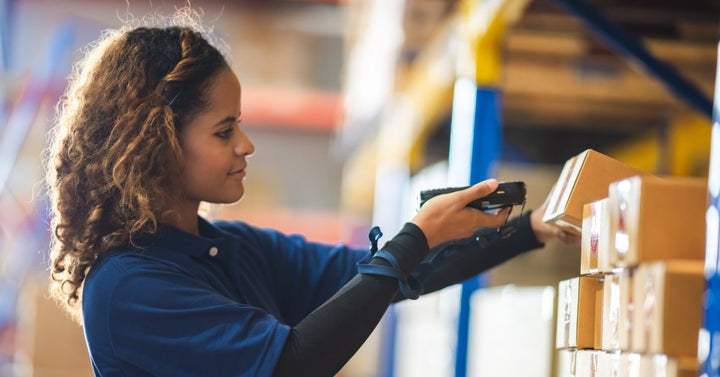 A young worker in a distribution center scanning inventory boxes that are stacked on an industrial shelf.