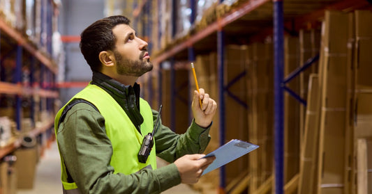 A warehouse worker is inspecting the shelves and storage system in the facility. He is holding a clipboard and a pencil.