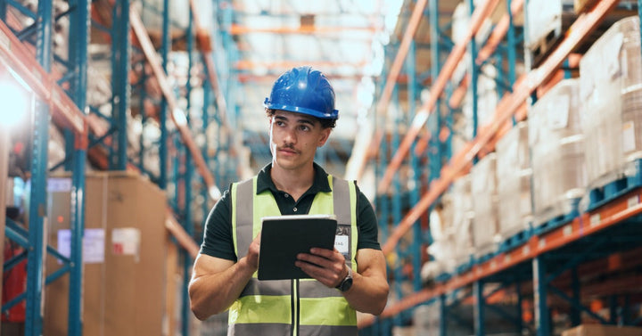 A man wearing a blue hard hat and yellow safety vest, holding a tablet between shelves of boxes in a warehouse.