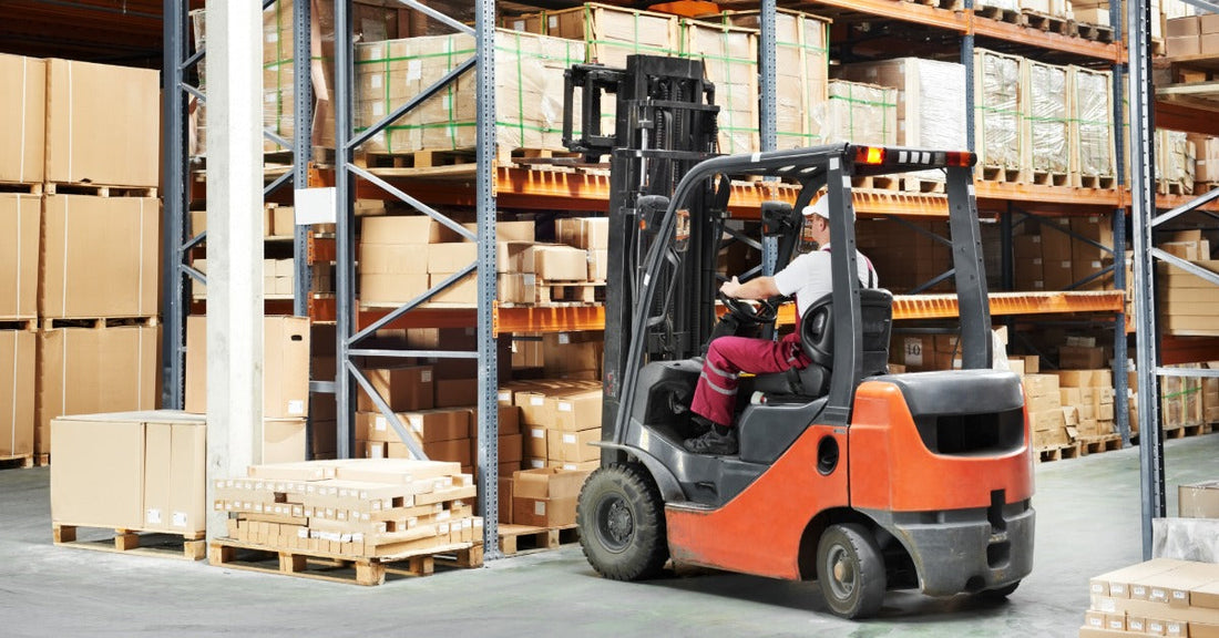 A worker driving an orange forklift through a warehouse aisle between tall racks of stacked boxes and pallets.