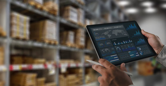 Hands holding a tablet with data charts and a stylus in a warehouse aisle with blurred shelves and stacked boxes.