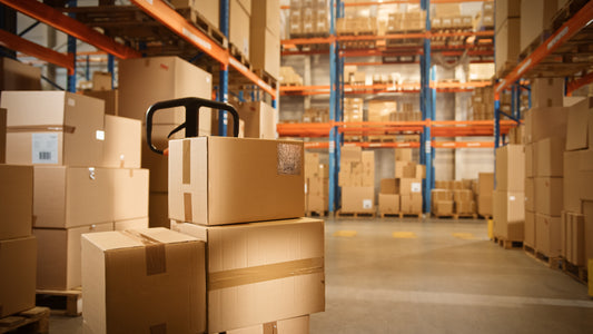 A manual pallet truck loaded with cardboard boxes sits in the middle of an aisle between pallet racks.