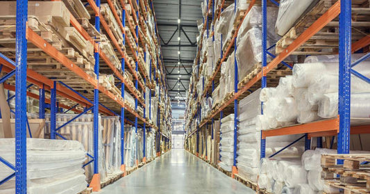 Two pallet racks and the clear aisle between them. The racks are mostly full and house various types of goods on wooden pallets.