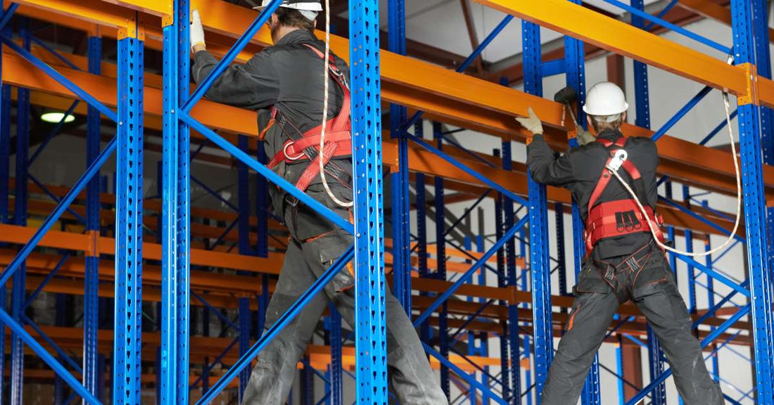 Two individuals wearing hard hats and harnesses installing a blue and orange pallet racking system in a warehouse.