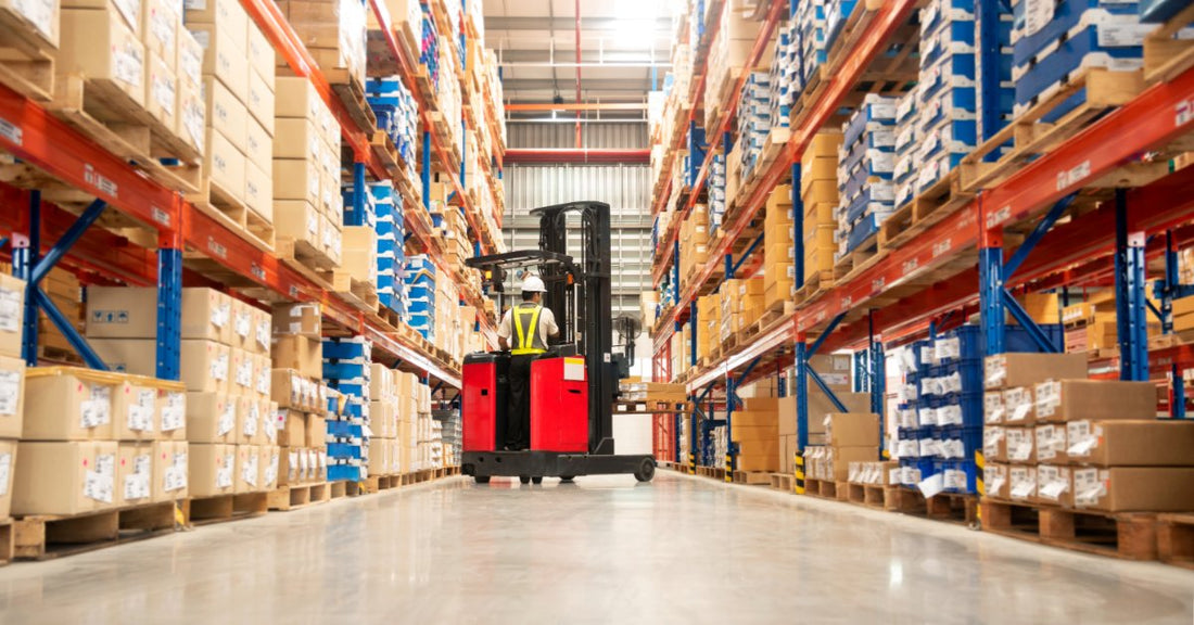 A worker driving a red forklift down a warehouse aisle between tall racks stacked with boxes and pallets.